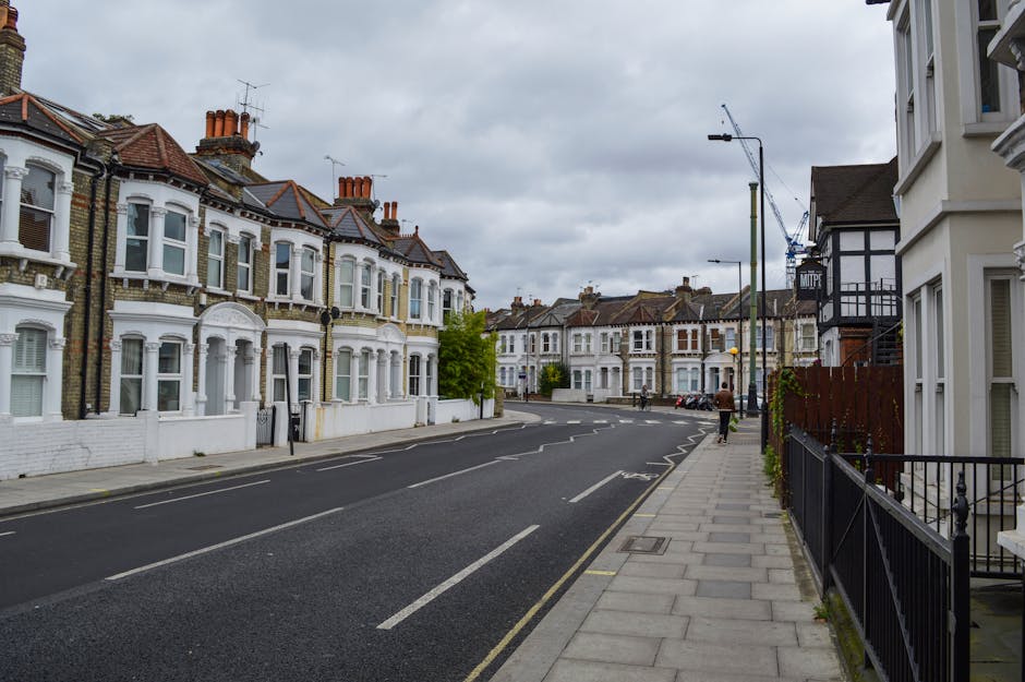 A street view of residential houses with Victorian-style architecture, featuring bay windows, brick facades, and terracotta chimneys, on a cloudy day. The pavement runs along the right side, with a bl