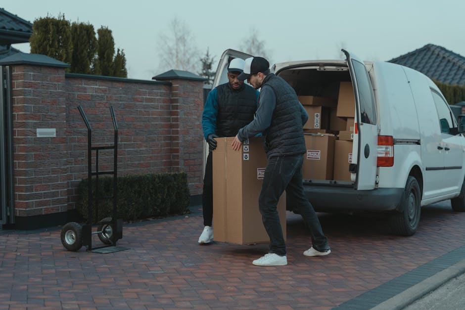 A man with a serious expression stands inside the open rear section of a commercial van during a home relocation process, with his arms crossed. The van's interior contains numerous cardboard boxes of
